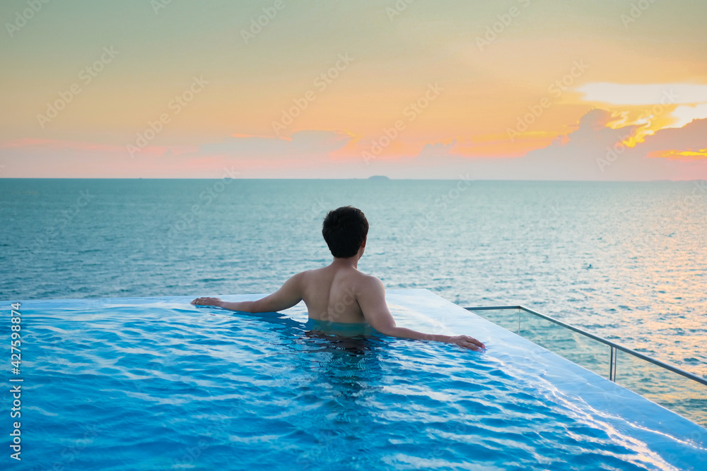 A man relaxing in a swimming pool at a luxury resort with blue sky and blue sea background. Young man enjoying beautiful sunset in luxury hotel near swimming pool. Calmness and serenity. 