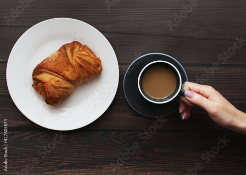 Fototapeta Naklejka Na Ścianę i Meble -  In the girl's hand a cup of coffee, a croissant, a roll. Delicious gourmet breakfast on wooden background. Flat lay, top view. Place for your text.
