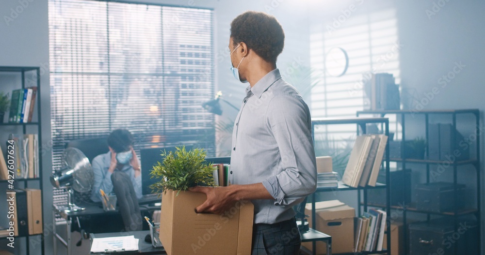 Portrait of young African American male worker in medical mask standing in cabinet holding carton box in hands with his stuff and looking at camera, fired from work, new employee at office Portrait of young African American male worker in medical mask standing in cabinet holding carton box in hands with his stuff and looking at camera, fired from work, new employee at office