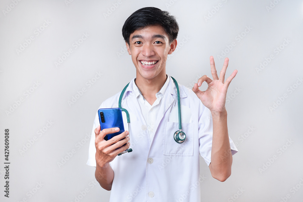 Happy Young Asian doctor smiling showing okay sign holding a mobile phone Happy Young Asian doctor smiling showing okay sign holding a mobile phone