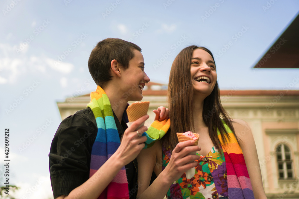 A young lesbian couple embraces their love. They are walking the streets and eating ice cream covered together with a rainbow colors scarf. A young lesbian couple embraces their love. They are walking the streets and eating ice cream covered together with a rainbow colors scarf.