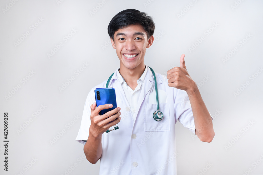 Friendly Young Asian doctor man smiling giving thumb up while holding a mobile phone Friendly Young Asian doctor man smiling giving thumb up while holding a mobile phone