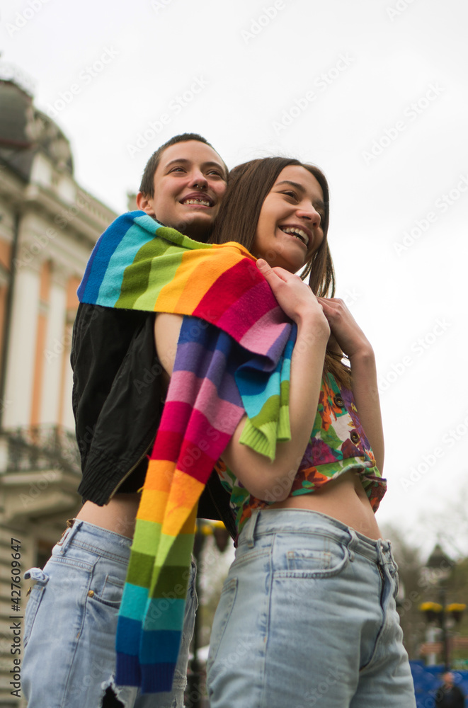 A young lesbian couple embraces their love. They are hugging on the street, covered together with a rainbow colors scarf.