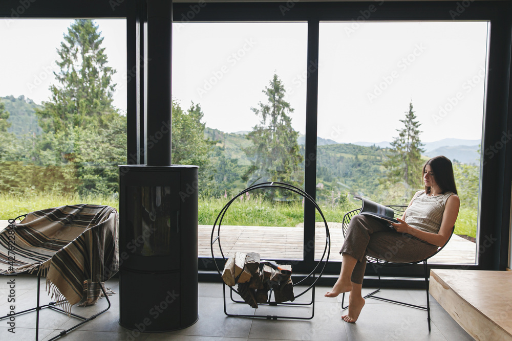 Beautiful stylish woman reading book, sitting on chair at fireplace on background of mountain hills