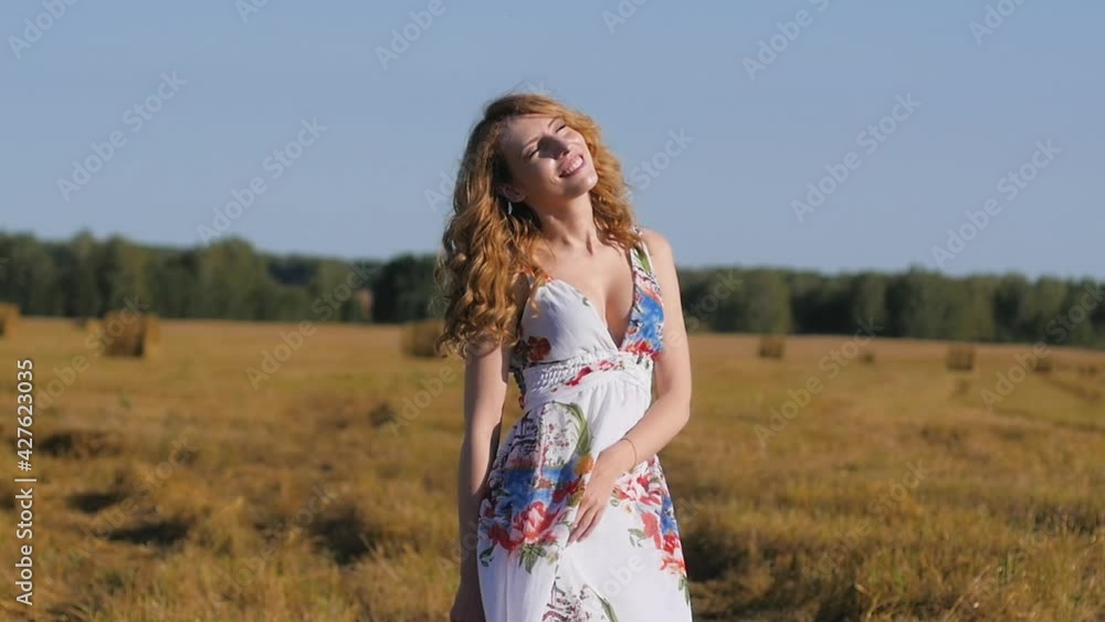 beautiful red-haired girl in a white dress and hat stands against the background of a field and haystacks. Young woman at sunset on the background of nature beautiful red-haired girl in a white dress and hat stands against the background of a field and haystacks. Young woman at sunset on the background of nature