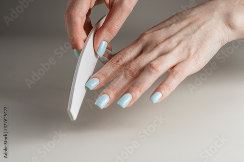 Woman hands with baby blue manicure  holding nail file on light beige background. Nail care and beauty concept.