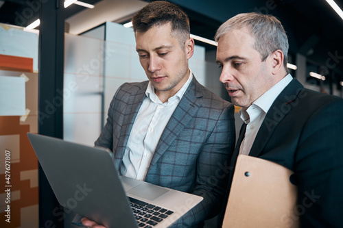 Two businessmen using modern laptop in office corridor