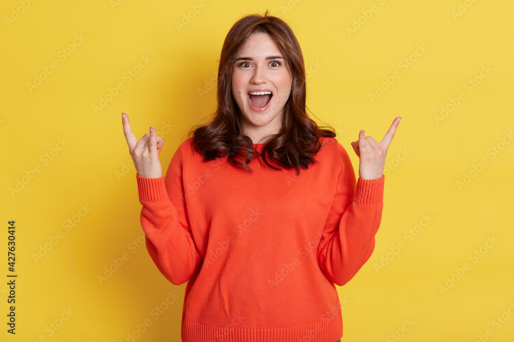 Φωτογραφία Excited dark haired female wearing casual sweater showing rock sing with both hands, exclaiming happily, looks at camera with positive expression, isolated over yellow background