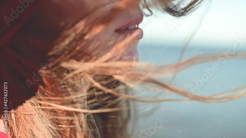 Close-up of a young Caucasian woman's hair fluttering in slow motion in the wind. Shallow depth of field. Girl in red dress and hat dreamer walk by the sea