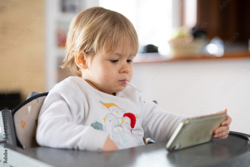 Cute little boy using smartphone, looking at screen, curious child holding phone in hands, sitting on a chair at home. Cute little boy using smartphone, looking at screen, curious child holding phone in hands, sitting on a chair at home.
