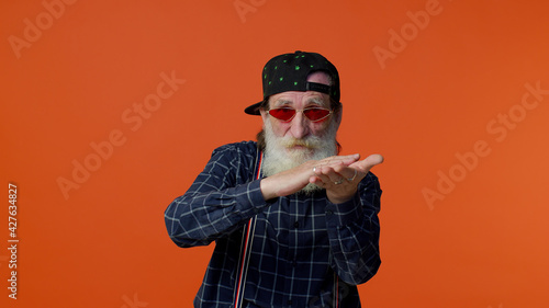 Mature old bearded grandfather in sunglasses and cap showing wasting or throwing money around hand gesture, more tips dreaming about big profit body language. Senior man isolated on orange background photo