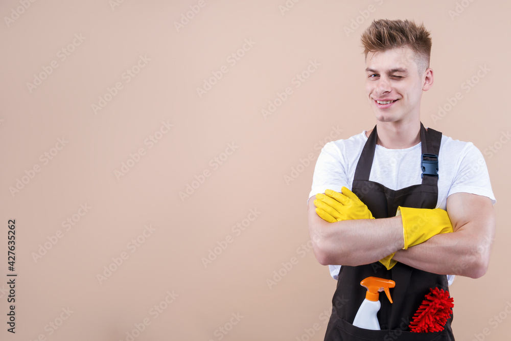 Young smiling man wearing cleaner apron and rubber yellow gloves isolated on beige background. Happy houseworker, househusband is ready for hard work, washing house. Professional cleaning service. Young smiling man wearing cleaner apron and rubber yellow gloves isolated on beige background. Happy houseworker, househusband is ready for hard work, washing house. Professional cleaning service.