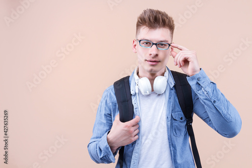 Portrait of smiling man wearing denim shirt, backpack standing against isolated on beige background. Young student is pointing spectacles with finger, thinking, focused on task. Back to school concept