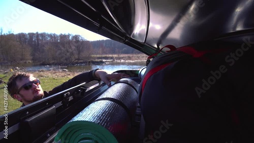 A man puts things in the roof rack of a car or in a cargo box, on a blurred background of a river and trees, on a summer day. family trip. Close-up Inside view.