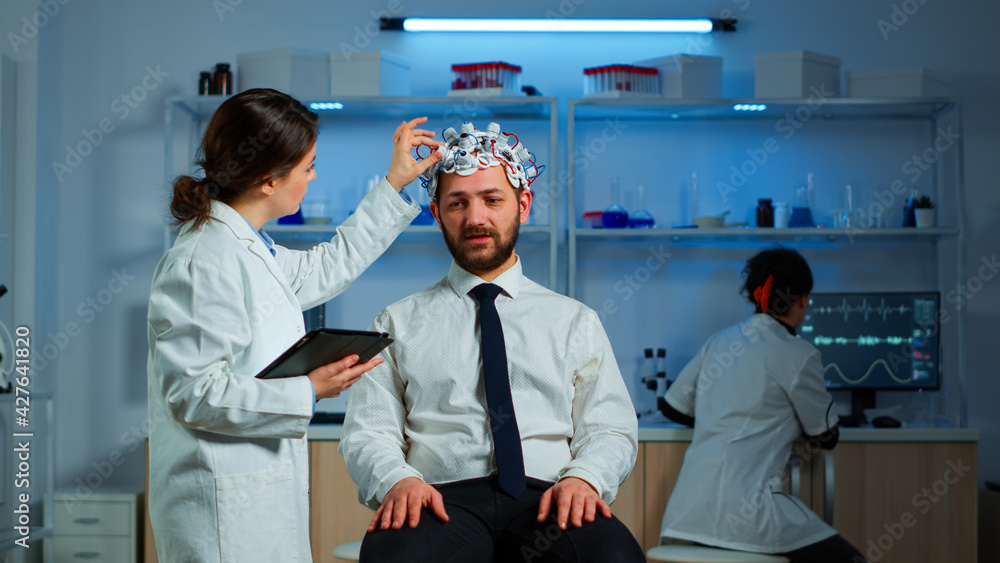 Obraz premium Young man wearing brainwave scanning headset discussing with neurological researcher about symptoms of disease. Medical scientist writing informations of brain scan on tablet working in laboratory