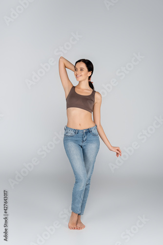 smiling woman in jeans and bra looking away while posing on grey