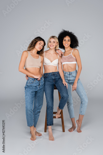 smiling multicultural women in bras and jeans posing near high stool on grey