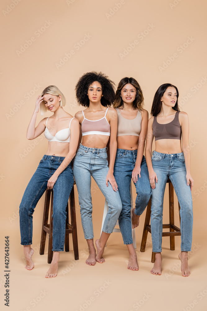 Fototapeta premium slim interracial women in bras and jeans posing near high stools on beige