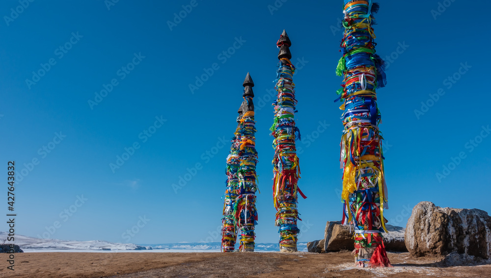 Naklejka premium Wooden tall pillars, standing in a row, are tied with multi-colored ritual ribbons. Nearby, on the ground, there are picturesque boulders. Clear blue sky. Mount Shamanka. Baikal