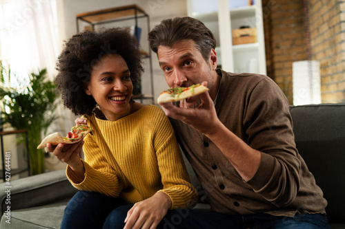 Cheerful young couple sitting on sofa at home. Happy woman and man eating pizza while watching a movie