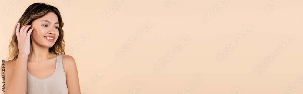 joyful asian woman fixing hair while looking away isolated on beige, banner joyful asian woman fixing hair while looking away isolated on beige, banner
