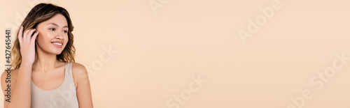 Wallpaper Mural joyful asian woman fixing hair while looking away isolated on beige, banner Torontodigital.ca