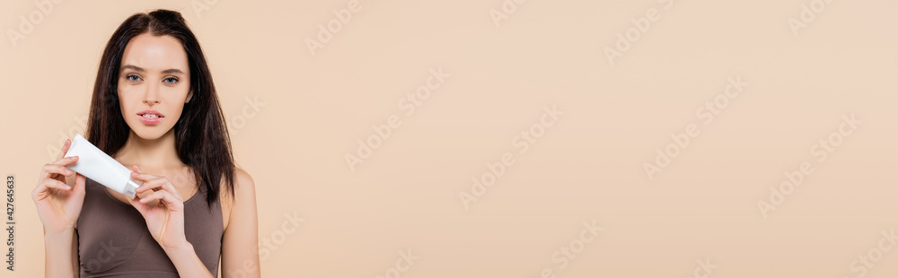 young woman looking at camera while holding cosmetic cream isolated on beige, banner young woman looking at camera while holding cosmetic cream isolated on beige, banner