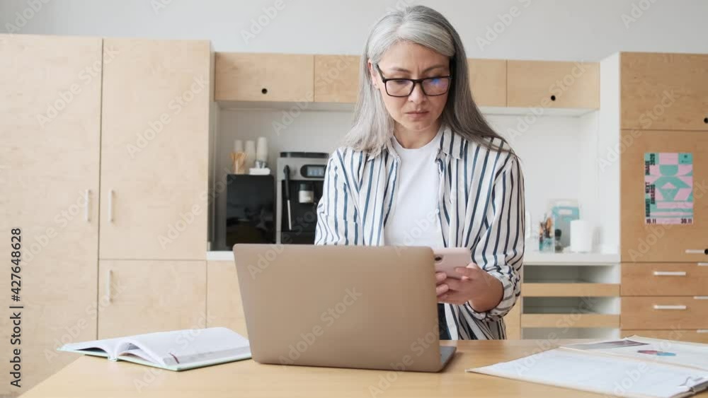 A serious elder business woman wearing eyeglasses is using her laptop computer while making notes in the kitchen at home A serious elder business woman wearing eyeglasses is using her laptop computer while making notes in the kitchen at home