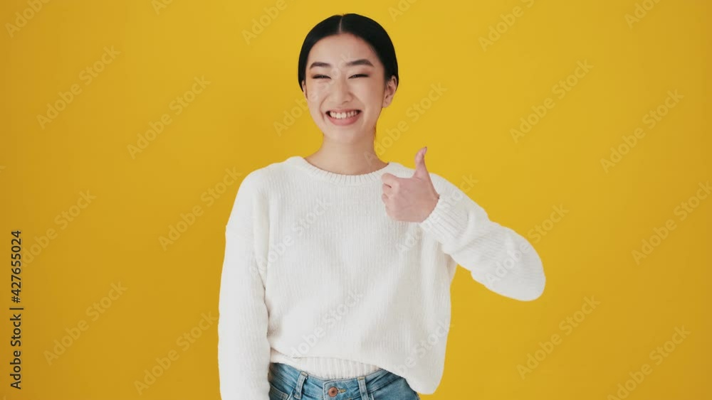 A smiling asian woman is doing thumb-up gesture standing isolated over yellow wall in the studio A smiling asian woman is doing thumb-up gesture standing isolated over yellow wall in the studio