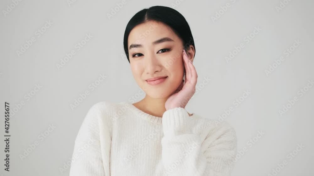 A charming asian woman wearing white sweater is posing to the camera standing isolated over white wall in the studio A charming asian woman wearing white sweater is posing to the camera standing isolated over white wall in the studio