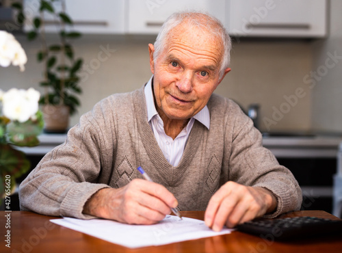 old gray haired man sits at table and writes on white sheet