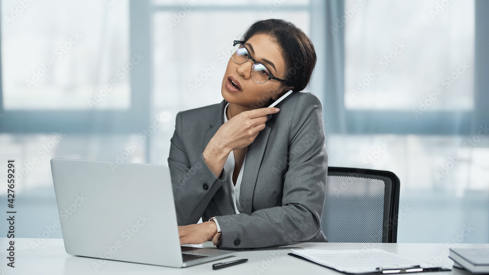 african american businesswoman talking on smartphone near laptop on desk african american businesswoman talking on smartphone near laptop on desk