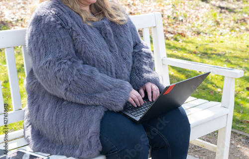 Frau in einer Kaninchenfell Jacke arbeitet im Garten mit einem Notebook im Homeoffice  