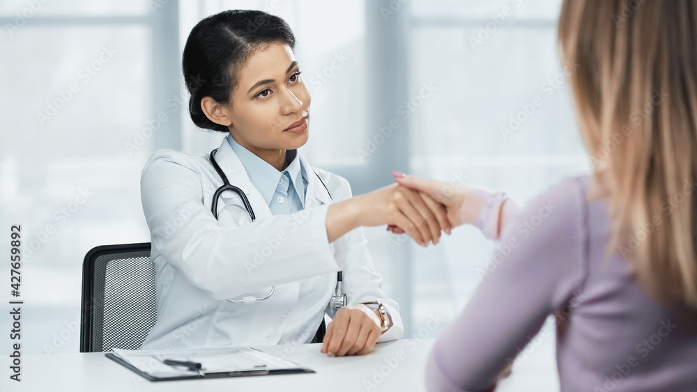 african american doctor in white coat shaking hands with patient on blurred foreground