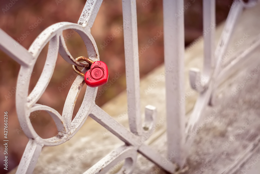 A small iron lock in the form of a red heart is chained to the fence of the bridge on a brown background. Wedding ceremony as a sign of loyalty and well-being for many years. The beloved ritual.