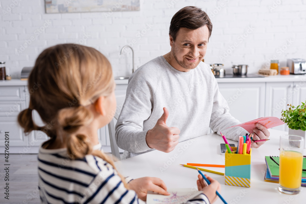 joyful man showing thumb up near daughter holding color pencil on blurred foreground joyful man showing thumb up near daughter holding color pencil on blurred foreground