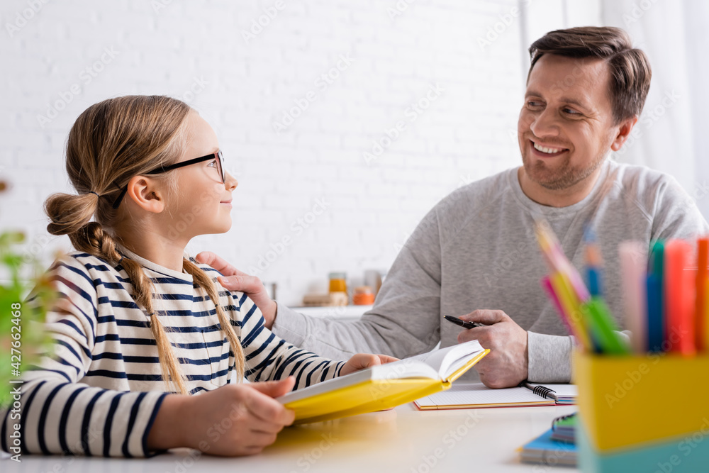 smiling man looking at daughter holding textbook while doing homework on blurred foreground smiling man looking at daughter holding textbook while doing homework on blurred foreground