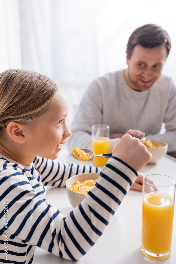 girl eating corn flakes near smiling father on blurred background girl eating corn flakes near smiling father on blurred background