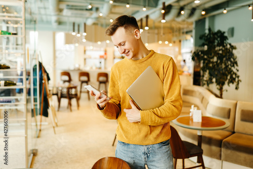 Young smiling business man freelancer in casual yellow sweater and jeans working remotely using laptop and phone sitting in cafe, selective focus