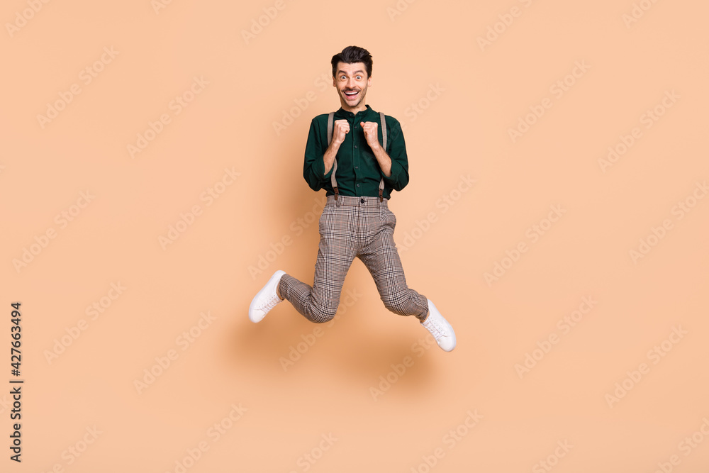 Full length photo of young man happy smile excited rejoice victory fists hands jump isolated over beige color background Full length photo of young man happy smile excited rejoice victory fists hands jump isolated over beige color background