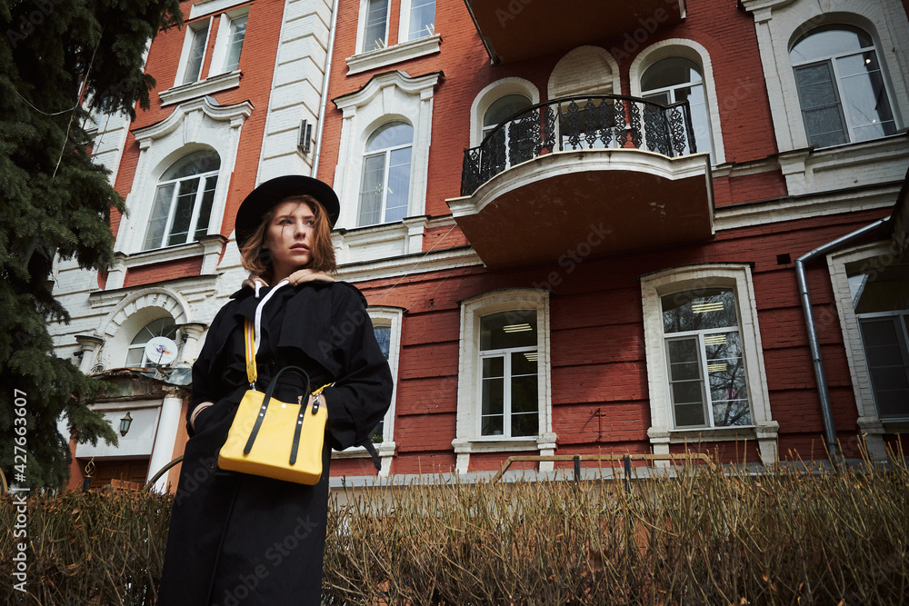 The girl gazes at the horizon against the background of a red building with a yellow bag in a black cloak