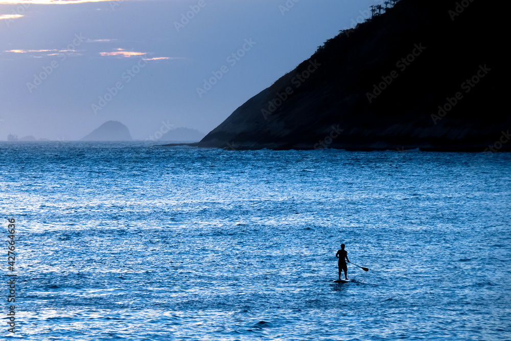 Person on stand up board in the blue expanse of the sea, in the late afternoon, with silhouette of Morro das Andorinhas, Itacoatiara Beach, Niteroi, State of Rio de Janeiro, Brazil