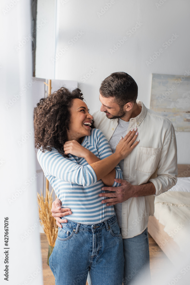joyful multiethnic couple laughing while hugging at home joyful multiethnic couple laughing while hugging at home