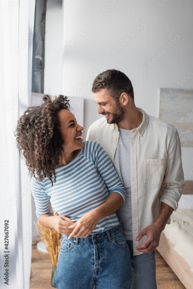 happy multiethnic couple laughing while hugging at home happy multiethnic couple laughing while hugging at home