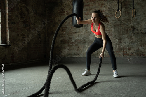Young fit sportswoman working out in fitness training gym. Woman exercising with battle ropes at gym