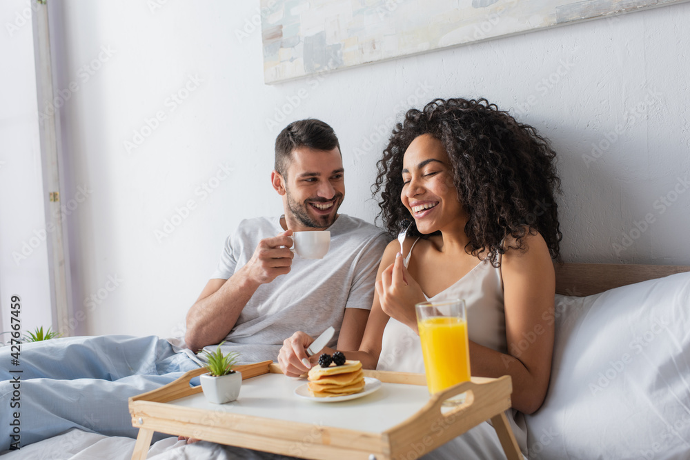 cheerful african american woman holding fork with blackberry near pancakes on tray and boyfriend with cup of coffee cheerful african american woman holding fork with blackberry near pancakes on tray and boyfriend with cup of coffee