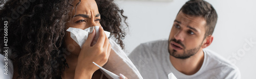 sick african american woman sneezing in napkin near worried boyfriend, banner