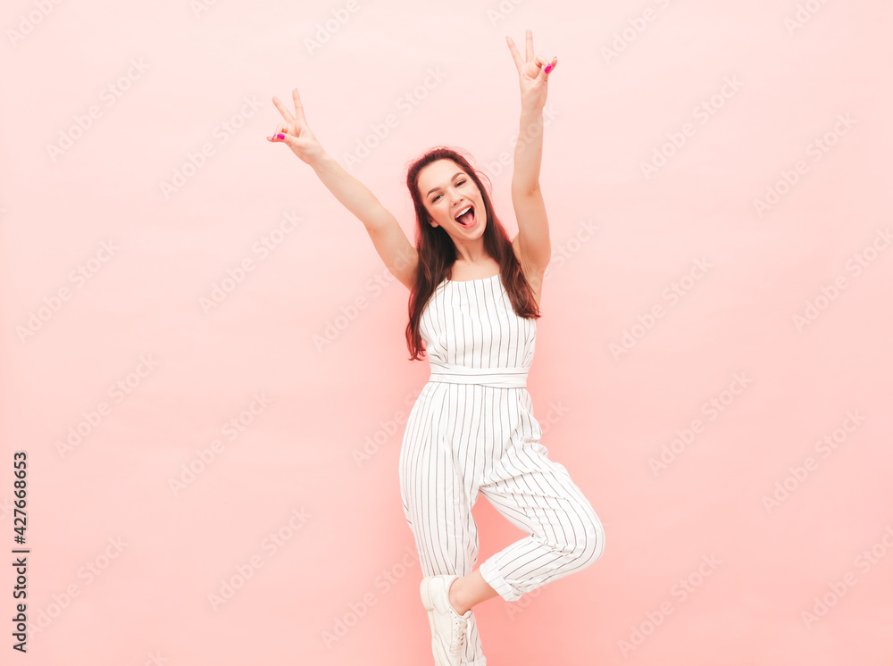 Portrait of young beautiful smiling female in trendy summer hipster overalls clothes. Sexy carefree woman posing near pink wall in studio. Positive model having fun indoors. Shows peace sign