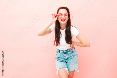 Young beautiful smiling female in trendy summer hipster clothes. Sexy carefree woman posing near pink wall in studio. Positive model having fun indoors. Cheerful and happy © halayalex