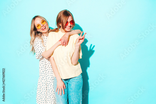 Two young beautiful blond smiling hipster female in trendy summer clothes. Sexy carefree women posing near blue wall in studio. Trendy and positive models having fun in sunglasses © halayalex